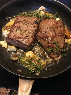 two steaks are being cooked in a skillet with garlic and herbs on the side