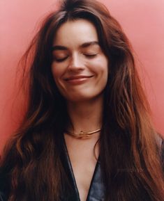 a woman with long hair smiling and wearing a choker necklace on her neck, in front of a pink wall