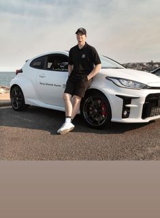 a man sitting on the hood of a white sports car next to the ocean with his legs crossed