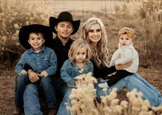 a family sitting on the ground in front of some tall grass and bushes with one child wearing a cowboy hat