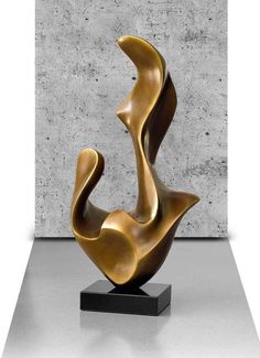 a bronze sculpture sitting on top of a black stand in front of a concrete wall