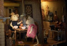 two mice sitting at a table eating food in a miniature kitchen area with lights on