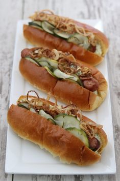 three hot dogs on buns with pickles, onions and other toppings sitting on a white plate