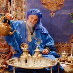 a man in blue sitting down and pouring water from a teapot on top of a table