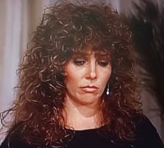 a woman with long curly hair standing in front of a tv screen and looking at the camera