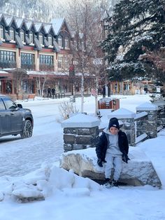 a young boy sitting on a rock in the middle of a snow covered street next to a building