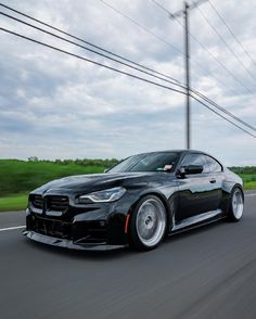 a black bmw is driving down the road with power lines in the background and clouds in the sky