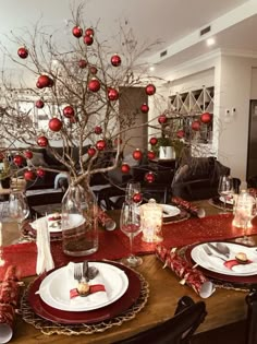 a dining room table set for christmas with red ornaments on the branches and place settings