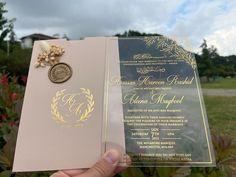 a person holding up a wedding card in front of some flowers and trees with clouds