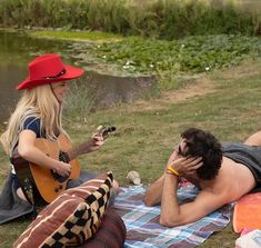 two people sitting on the grass playing guitar and singing to someone else with a red hat