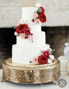 a white wedding cake with red and pink flowers on the top is sitting on a gold stand