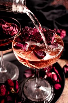 two glasses filled with liquid and rose petals on top of a wooden table next to each other