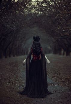 a woman in a black dress with a crown on her head is walking through the woods