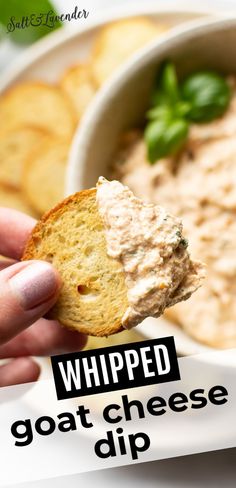 a hand holding a cracker over a bowl of dip with basil on top and the words whipped goat cheese dip above it