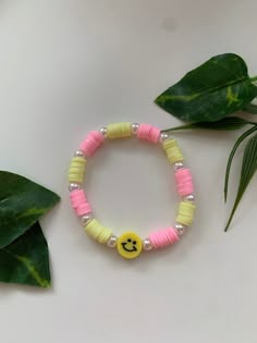 a yellow and pink beaded bracelet with a smiley face charm on it next to a green plant