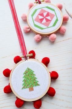 two cross stitch christmas ornaments hanging from red ribbon on white wooden table with pink pom poms