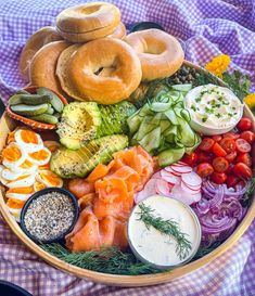 an assortment of food in a wooden bowl on a purple checkered table cloth, including bagels, tomatoes, cucumbers, onions and other vegetables