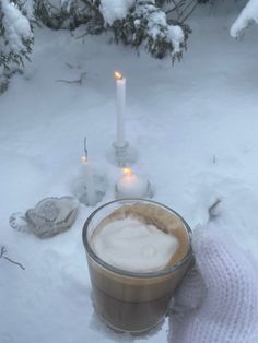 a cup of coffee sitting on top of snow covered ground next to two lit candles