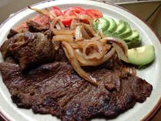 a white plate topped with steak, onions and sliced avocado on top of a wooden table