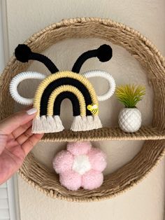 a hand holding a small knitted bee on top of a shelf next to a potted plant