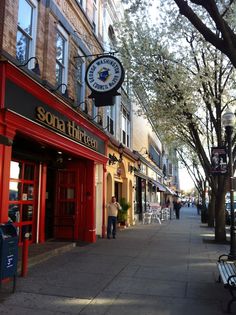 the sidewalk is lined with shops and people walking down it, along with trees on both sides