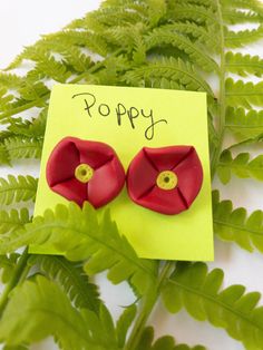 a pair of red flower earrings sitting on top of a green leafy plant next to a yellow post - it note