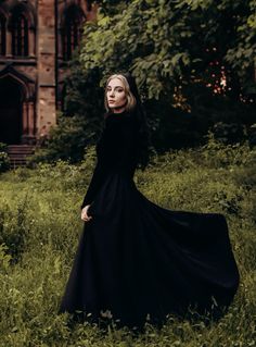 a woman in a long black dress is standing in the grass near a building and trees