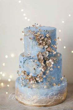 a three tiered blue cake with flowers on it's side and lights in the background