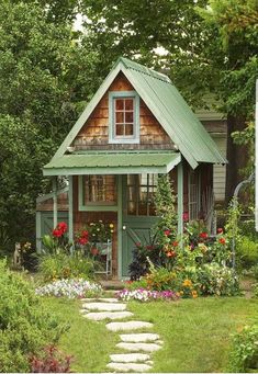 a small green house surrounded by flowers and greenery in the yard with stepping stones leading up to it