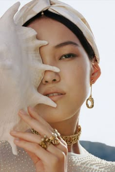 a woman holding a seashell in front of her face while wearing gold jewelry and earrings