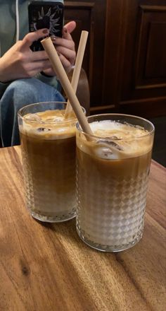 two glasses filled with liquid sitting on top of a wooden table next to a person holding a cell phone