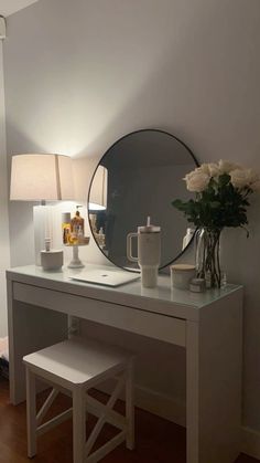 a white desk with a mirror, stool and vase on it next to a lamp