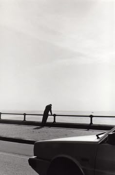 black and white photograph of man walking on the side of road next to car with ocean in background