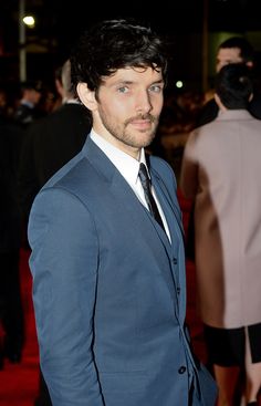 a man in a suit and tie standing on a red carpet with other people behind him