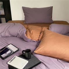 an unmade bed with purple sheets, pillows and a coffee cup on the end