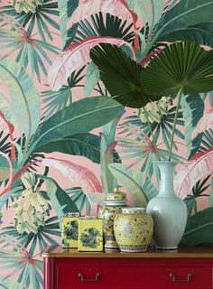 a red dresser topped with vases next to a wall covered in green leaves and flowers