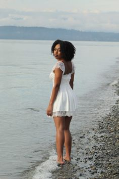 a woman is standing on the beach by the water wearing a white dress and sandals