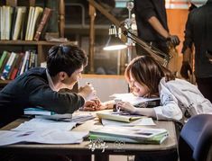 two people sitting at a table with books and papers on it, one person is writing