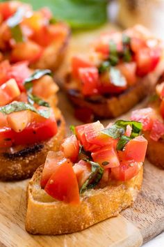 small pieces of bread with tomatoes and basil on top sitting on a wooden cutting board