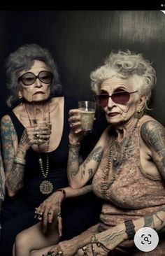 two older women sitting next to each other with tattoos on their arms, holding drinks