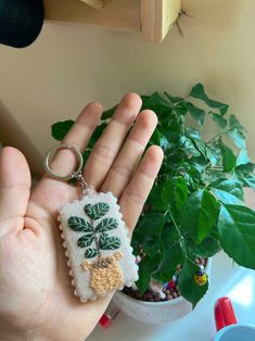 a hand holding a small keychain with a plant in the corner behind it