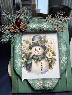 a green frame with a snowman on it and pine cones in the corner next to it