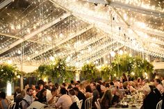 a large group of people sitting at tables in a room with lights hanging from the ceiling