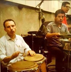 two men are playing drums in front of a microphone and another man is sitting behind them