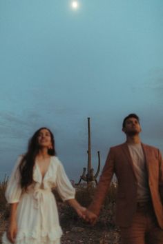 a man and woman holding hands in front of a cactus at night with the moon behind them