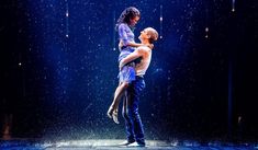 a man and woman dancing in the rain on stage with lights shining down behind them