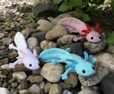 two crocheted geckos are laying on some rocks and gravel, one is pink and the other is blue