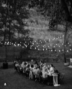 a group of people sitting around a table with lights strung from the trees in the background