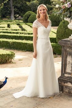 a woman in a white dress standing next to a peacock on a stone walkway with hedges and bushes behind her