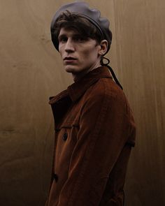 a man wearing a brown jacket and hat standing in front of a wooden wall with his hands on his hips